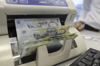 Bancrete de culoare galbenă și albastră trec printr-o mașină de numărat bani lângă un tastatură de calculator.