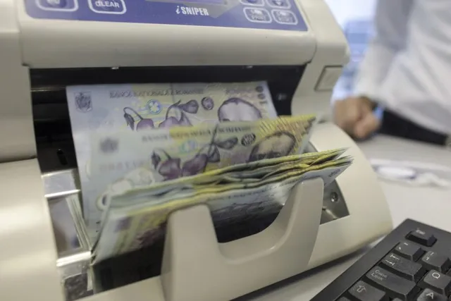 Bancrete de culoare galbenă și albastră trec printr-o mașină de numărat bani lângă un tastatură de calculator.