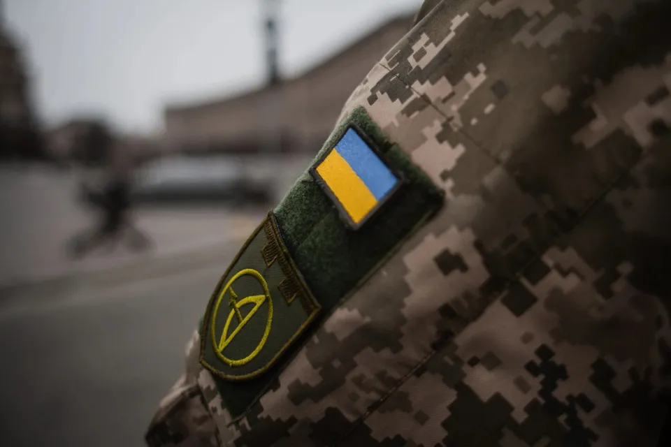 Un patch cu steagul ucrainean și un emblema circulară sunt cusute pe o uniformă militară cu model de camuflaj digital.