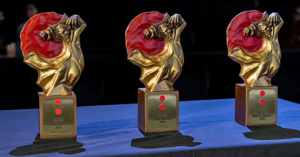 Trei trofee aurii cu o figură umană și o formă roșie sunt așezate pe un suport dreptunghiular pe o suprafață albastră.