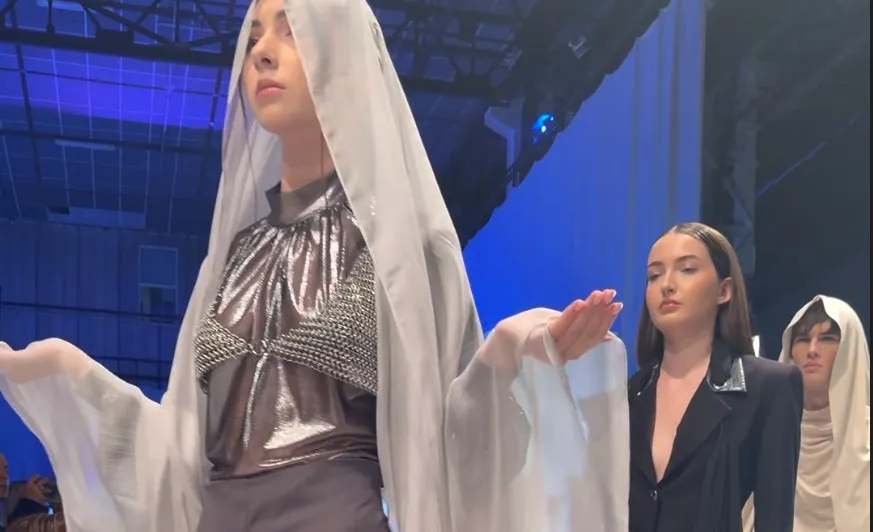 O femeie poartă o rochie metalizată și un voal alb lung, în timp ce alte două femei stau în spatele ei pe un podium.
