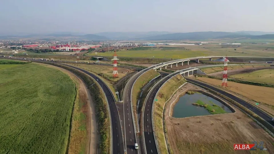 O vedere aeriană prezintă mai multe benzi de autostradă care se ramifică într-un nod rutier înconjurat de câmpuri verzi și dealuri îndepărtate.