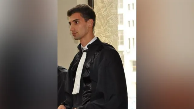 Un bărbat tânăr cu părul scurt și întunecat poartă o robă neagră peste o cămașă albă cu un papion negru.