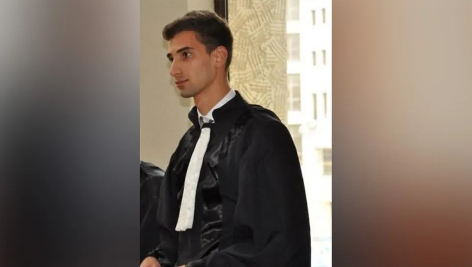 Un bărbat tânăr cu părul scurt și întunecat poartă o robă neagră peste o cămașă albă cu un papion negru.