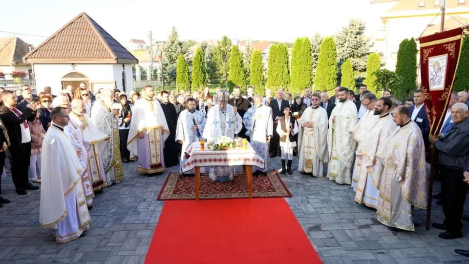 Un grup de oameni îmbrăcați în haine albe și liturgice stau în jurul unei mese acoperite cu un șorf, pe un covor roșu.