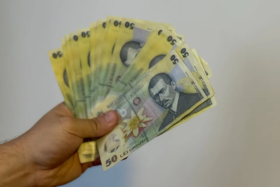 O mână ține un evantai de bancnote galbene de cincizeci de lei românești în fața unui fundal alb.