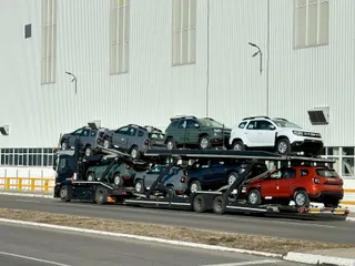 Un camion transportor de mașini transportă mai multe autoturisme de diferite culori și modele pe un drum lângă o clădire albă înaltă.