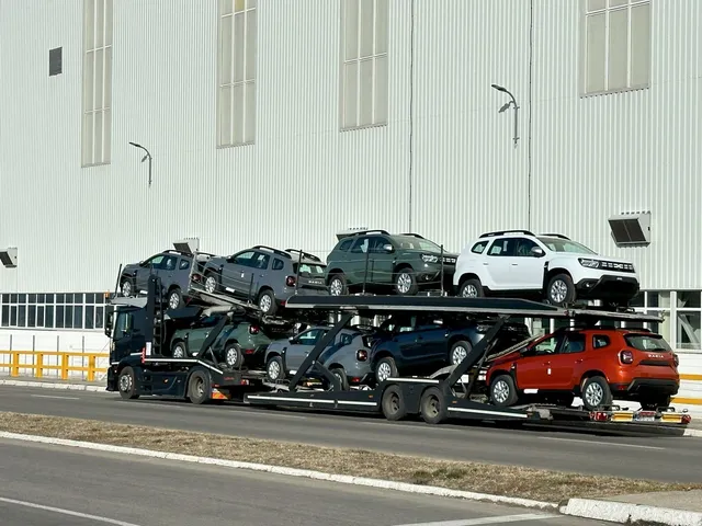 Un camion transportor de mașini transportă mai multe autoturisme de diferite culori și modele pe un drum lângă o clădire albă înaltă.
