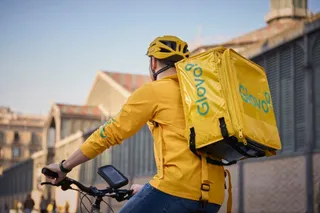 Un bărbat cu cască galbenă și tricou galben poartă un rucsac termosac galben pe spate în timp ce conduce o bicicletă.