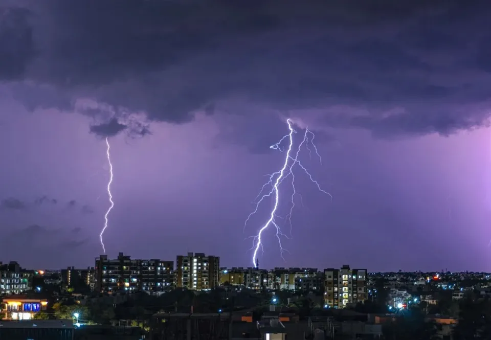 Mai multe fulgere străbat cerul închis deasupra unor clădiri urbane iluminate pe timp de noapte.