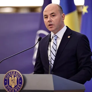 Un bărbat cu pielea deschisă, purtând un costum închis la culoare și o cravată cu dungi, stă în spatele unui podium cu emblema Guvernului României.
