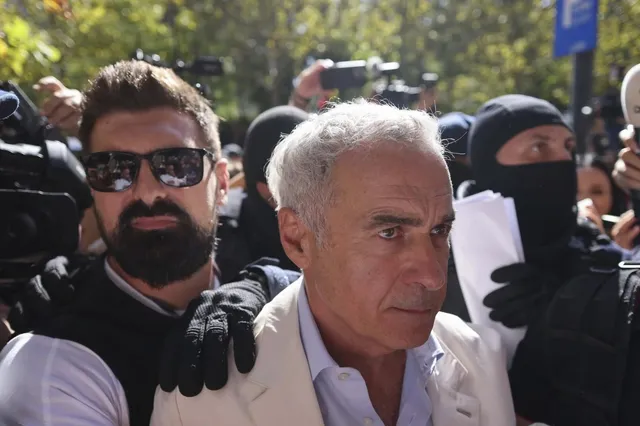 Un bărbat cu părul cărunt și sacou deschis este înconjurat de persoane, dintre care unul poartă ochelari de soare și o altul o mască neagră.