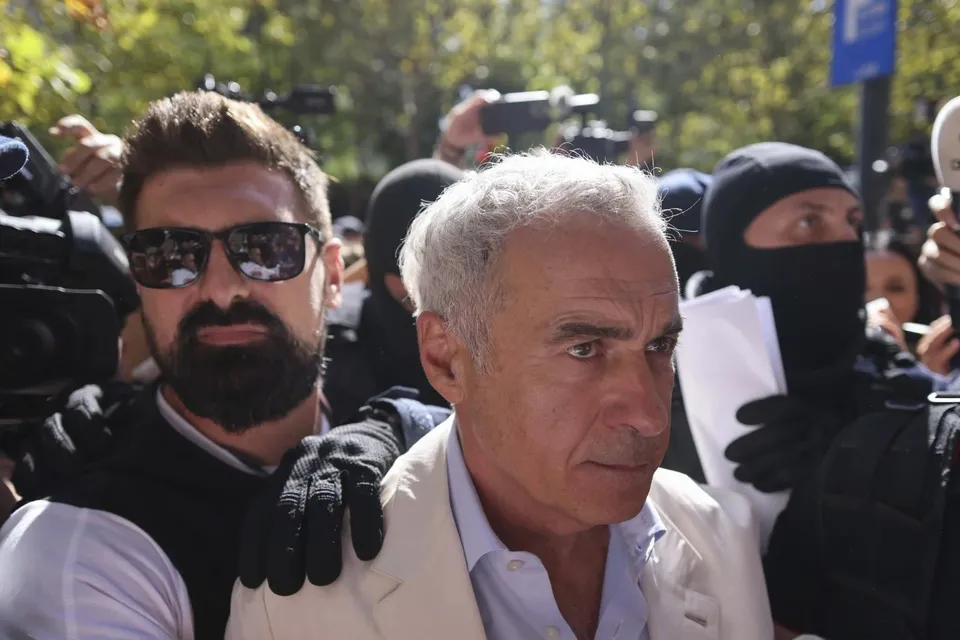 Un bărbat cu părul cărunt și sacou deschis este înconjurat de persoane, dintre care unul poartă ochelari de soare și o altul o mască neagră.
