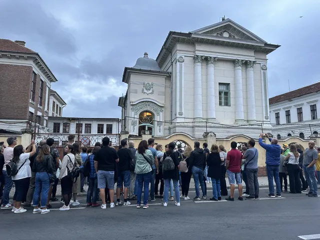 Un grup de persoane stă în picioare pe o stradă, privind spre o clădire albă cu coloane și un arc de intrare.