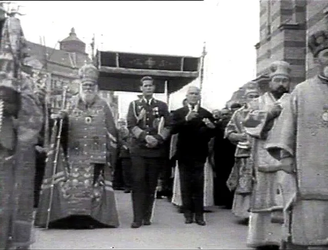 Un grup de bărbați în uniforme militare și haine religioase ceremoniale merge pe o stradă lângă o clădire cu fațadă striată.