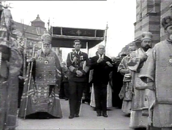 Un grup de bărbați în uniforme militare și haine religioase ceremoniale merge pe o stradă lângă o clădire cu fațadă striată.