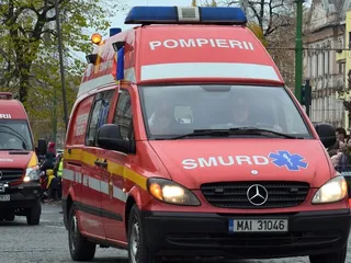 Un vehicul de serviciu roșu cu inscripția POMPIERII și SMURD este parcat pe un drum cu copaci în fundal.
