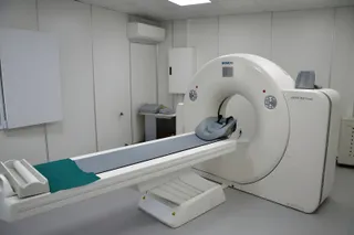 Un aparat medical de scanare de culoare albă este poziționat lângă un pat de examinare cu o suprafață gri și un accesoriu verde.