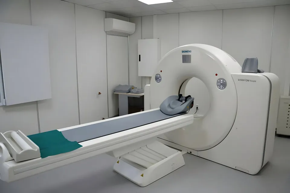 Un aparat medical de scanare de culoare albă este poziționat lângă un pat de examinare cu o suprafață gri și un accesoriu verde.