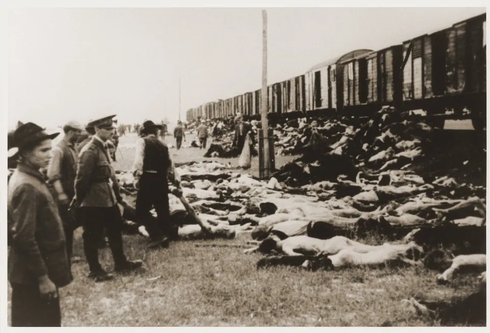 O fotografie alb-negru prezintă numeroase cadavre întinse pe sol lângă un tren lung format din vagoane de marfă, în prezența unor bărbați în uniformă.