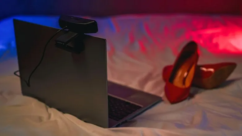 Un laptop cu o cameră web atașată stă deschis pe un pat lângă o pereche de pantofi cu toc roșii sub lumini colorate.
