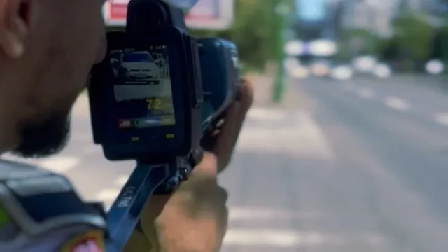 O persoană ține un aparat de măsurat viteza orientat spre o stradă cu mașini circulând în fundalul neclar.