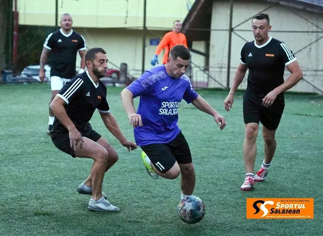 Patru bărbați joacă un meci de fotbal pe un teren verde, fiind un jucător în tricou albastru în centrul acțiunii.