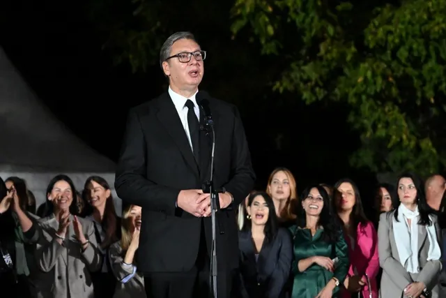Un bărbat cu ochelari și costum negru stă în fața unui microfon, în timp ce un grup de persoane îl privește în jur.