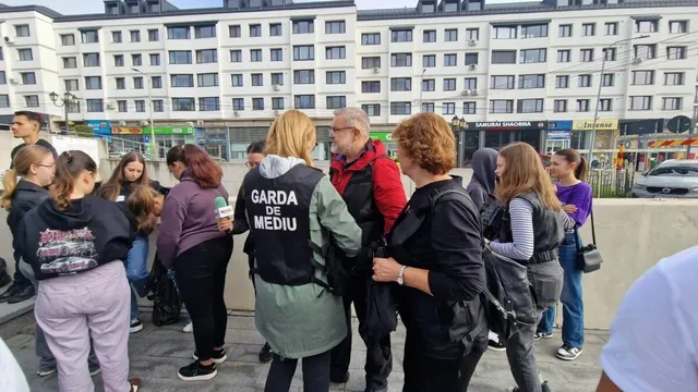 Un grup de persoane stă adunat pe un trotuar în fața unor clădiri de apartamente, purtând haine casual și vestă neagră cu text alb.
