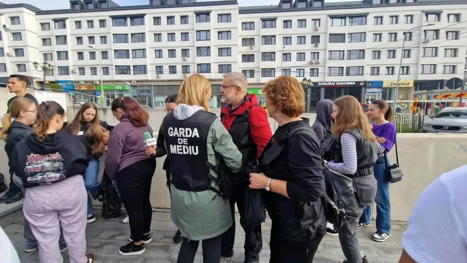 Un grup de persoane stă adunat pe un trotuar în fața unor clădiri de apartamente, purtând haine casual și vestă neagră cu text alb.