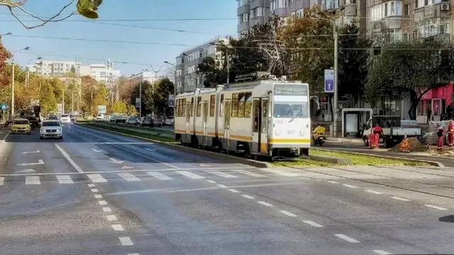 Un tramvai alb cu detalii galbene circulă pe șine într-o zonă urbană cu clădiri înalte și mașini pe carosabil.