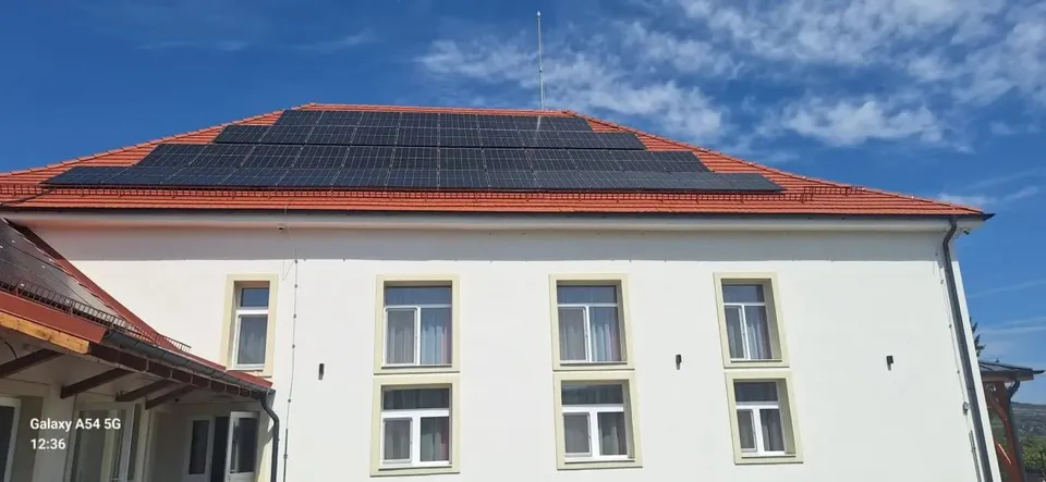 O clădire albă cu acoperiș de țiglă portocalie și panouri solare instalate pe partea superioară, sub un cer albastru cu nori.