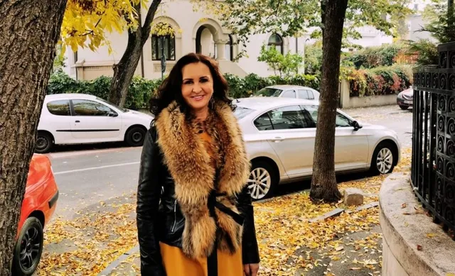 O femeie cu părul închis la culoare, purtând o jachetă neagră și o pălărie de blană maro, stă pe un trotuar acoperit de frunze galbene.