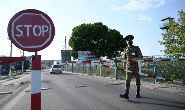 Un bărbat în uniformă militară și mască neagră stă lângă o barieră de drum marcată cu benzi albe și roșii, lângă un semn de stop roșu.