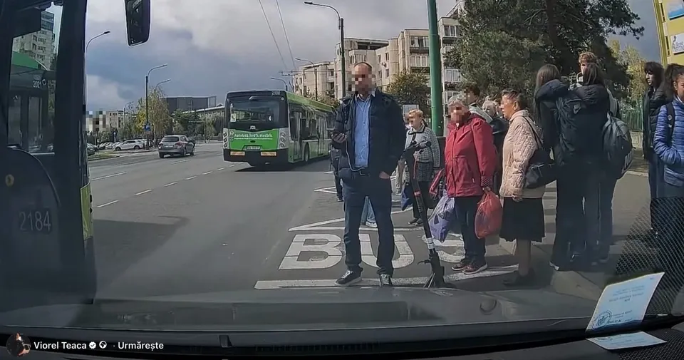 Un bărbat cu sacou și pantaloni închiși stă pe trotuar lângă un grup de oameni așteptând lângă un autobuz verde și alb pe o stradă.