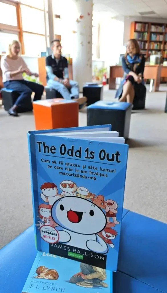 O carte cu copertă albastră intitulată "The Odd 1s Out" stă pe o suprafață albastră în prim-plan, cu trei persoane așezate pe cuburi în fundal.