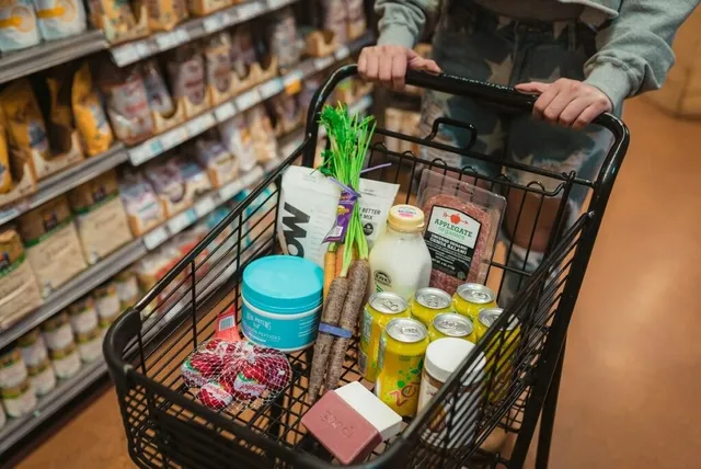 O persoană ține de mânerul unui cărucior de cumpărături umplut cu diverse produse alimentare, precum lapte, legume și conserve, într-un magazin.