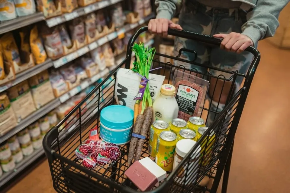 O persoană ține de mânerul unui cărucior de cumpărături umplut cu diverse produse alimentare, precum lapte, legume și conserve, într-un magazin.
