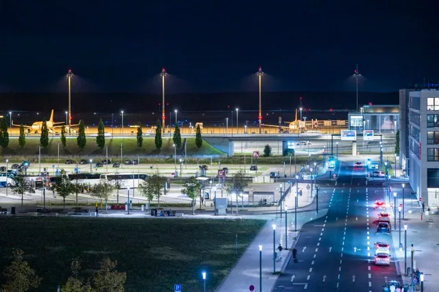 O vedere de noapte asupra unui aeroport cu avioane pe pistă, clădiri și o stradă cu mașini circulând sub lumină artificială.
