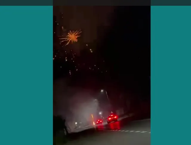 Artificii aprinse în cerul nopții deasupra unui drum pe lângă care circulă două mașini cu farurile aprinse.
