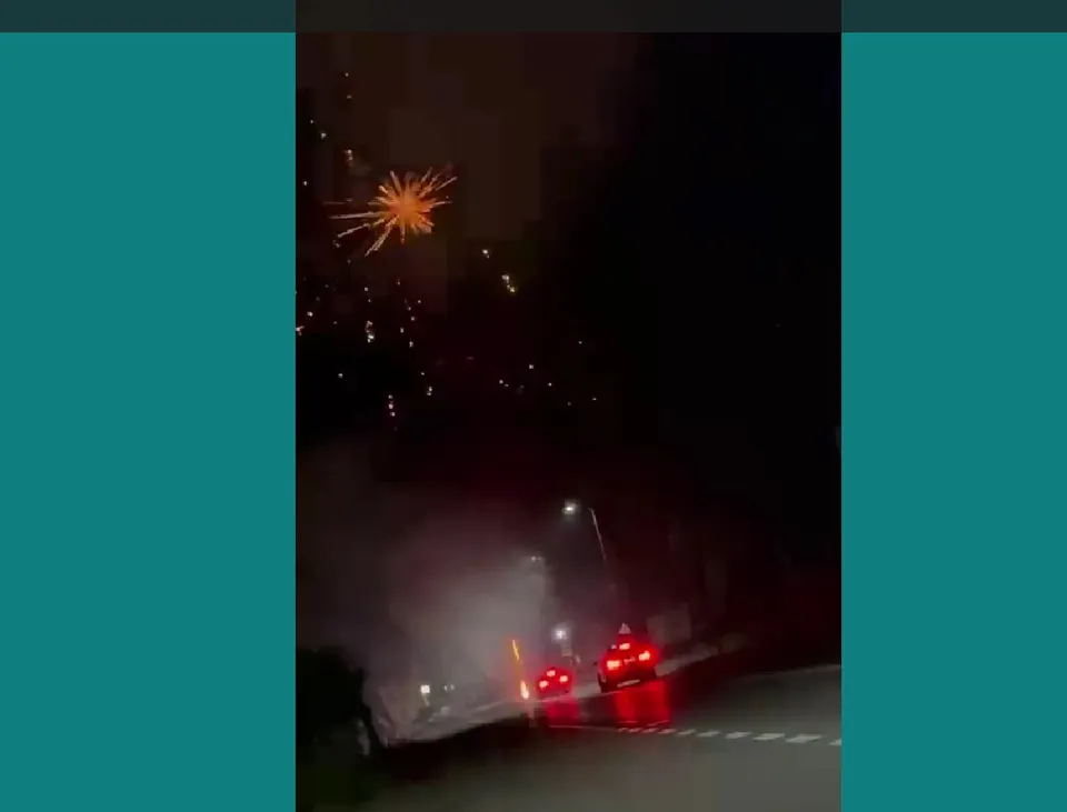 Artificii aprinse în cerul nopții deasupra unui drum pe lângă care circulă două mașini cu farurile aprinse.