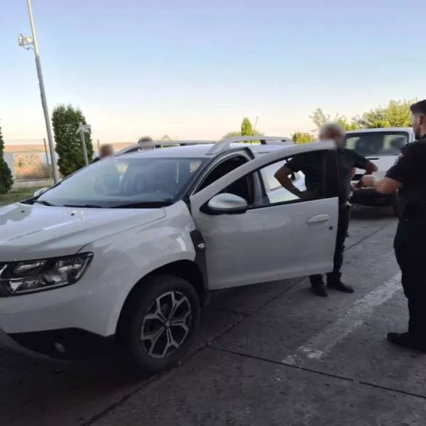 Un autoturism alb cu portiera șoferului deschisă este inspectat de doi bărbați în uniformă pe un teren asfaltat.