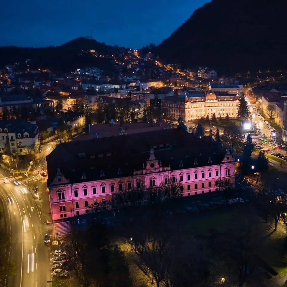 O clădire mare cu iluminare roz este situată lângă o stradă luminată, sub un deal cu case și lumini de oraș sub un cer închis.