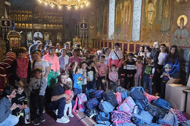 Un grup de copii și adulți stau într-o încăpere decorată cu picturi religioase, lângă un teanc de rucsacuri colorate pe podea.