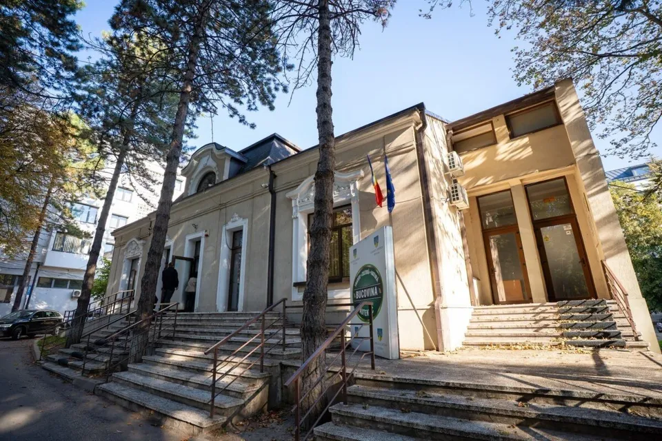 O clădire cu fațadă deschisă, trepte de beton și copaci înalți în jurul acesteia, sub un cer senin.