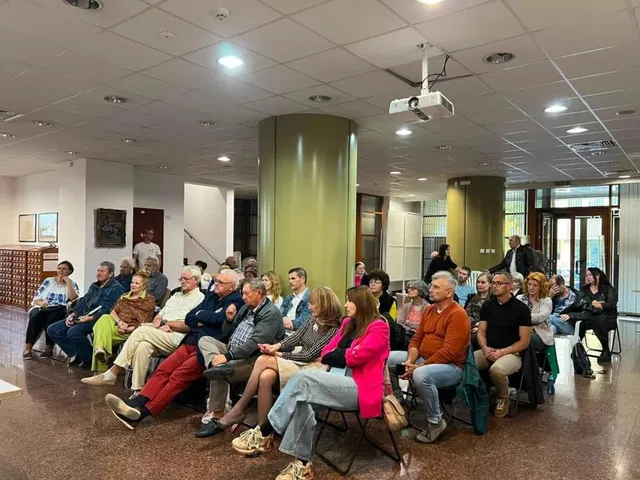 Un grup de persoane stă așezat pe scaune într-o încăpere interioară cu tavan cu panouri și un proiector suspendat.