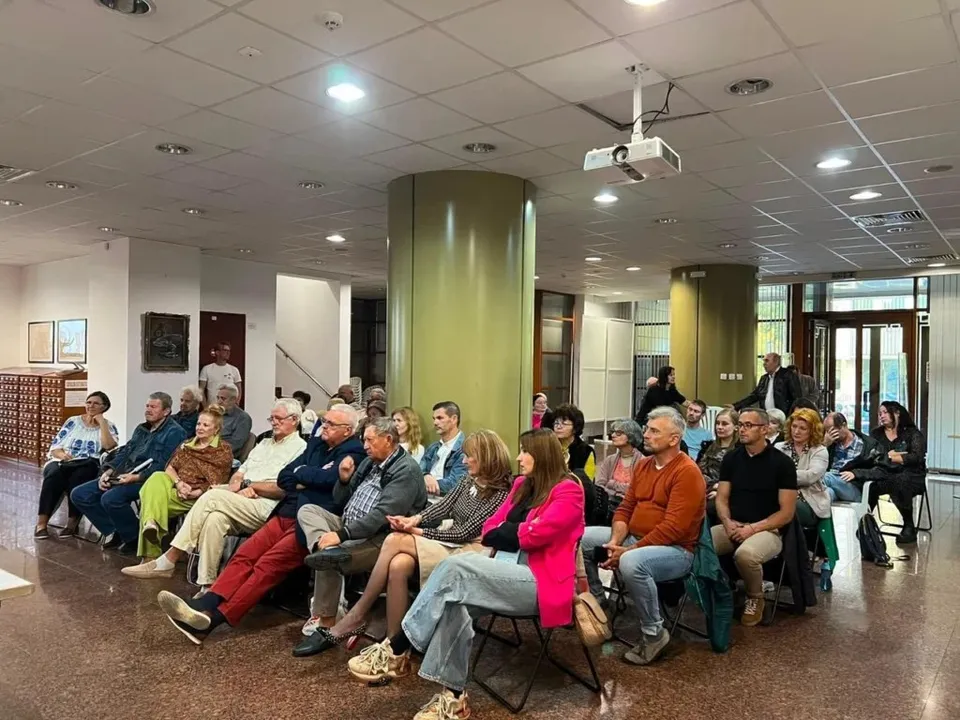 Un grup de persoane stă așezat pe scaune într-o încăpere interioară cu tavan cu panouri și un proiector suspendat.