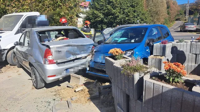 Două autoturisme, unul gri și unul albastru, sunt implicate într-un accident pe un drum lângă un gard de beton cu flori.
