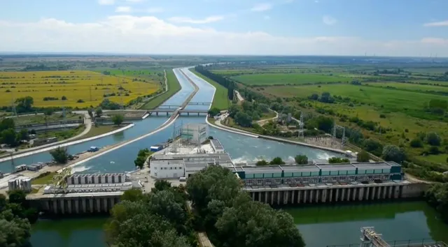 O vedere aeriană prezintă un canal de apă cu o barajă, vegetație verde și câmpuri agricole sub un cer înnorat.