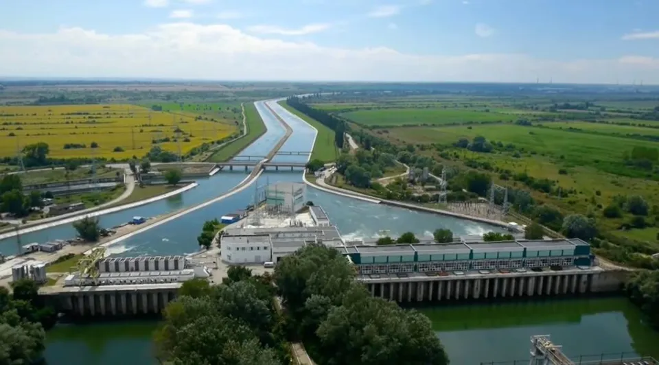 O vedere aeriană prezintă un canal de apă cu o barajă, vegetație verde și câmpuri agricole sub un cer înnorat.